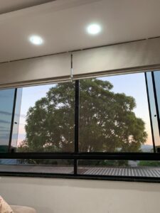 Imagem de janela de folhas de vidro deslizante com tela mosquiteira deslizante dupla com árvore ao fundo, pôr do sol, com iluminação interna. É possível ver a barra de uma cortina rolô na parte superior da janela.