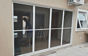 Ambiente fechado por uma tela mosquiteira porta camarão de quatro folhas. É possível enxergar o vulto de uma escada e um trabalhador.